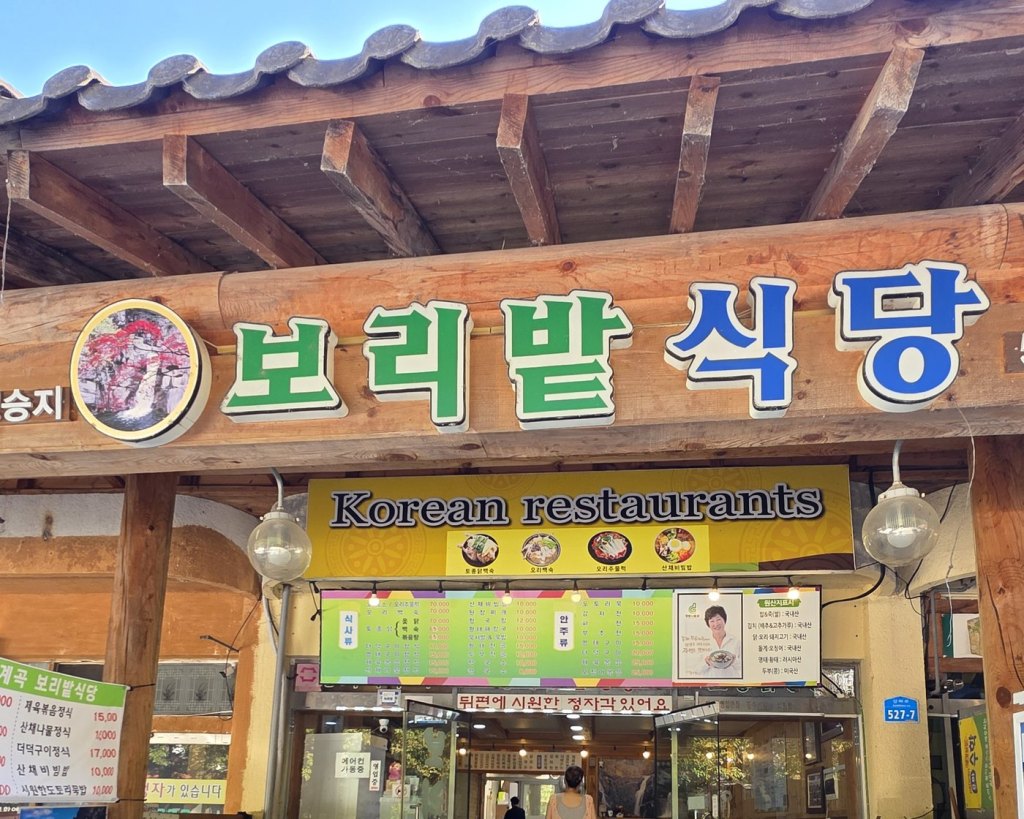 나무 기둥 위 「보리밭 식당」 간판과 그 아래 메뉴판이 보이는 식당 입구