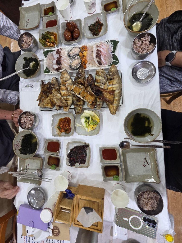 흰 식탁보 위에 생선구이, 모듬회, 밑반찬이 여러 접시 차려진 저녁상